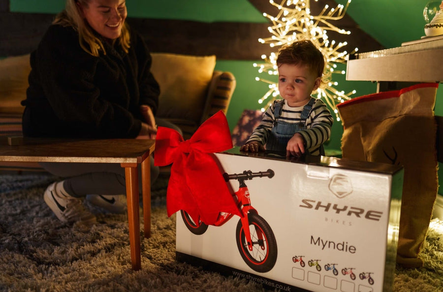 A child in overalls sits behind a large white product box for a balance bike, tied with a red bow, while an adult kneels beside it in a cozy indoor setting with warm lighting and a Christmas tree in the background. The box shows the brand name SHYRE and model name Myndie.