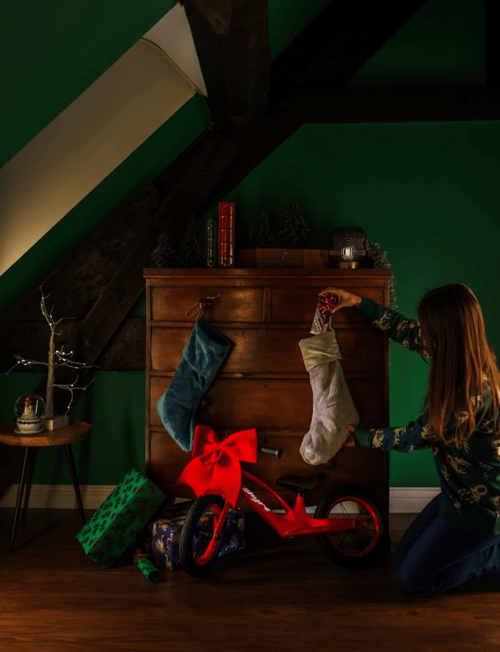 A woman kneels beside a red balance bike in a decorated attic room, hanging a white stocking on a wooden dresser while another stocking hangs on a drawer above. A wrapped gift box, books, and small holiday decor are visible against dark green walls.