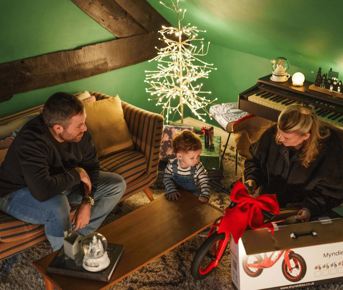 A family of three gathers in a cozy room with green walls and wooden beams, celebrating around a gift box for a red children's bicycle. A child is in the center, and an adult man and woman are seated on either side, smiling and interacting.